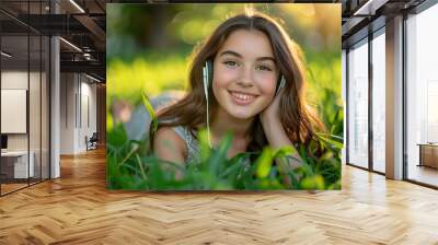 close-up portrait of a young woman lying on lush green grass, smiling and listening to music with headphones on a sunny spring day. Wall mural