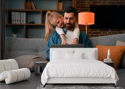 Cute active naughty girl noisy child plays with daddy hugs father distract parental attention disturbs parent from work on laptop at home single loving caucasian dad man calms down restless daughter Wall mural