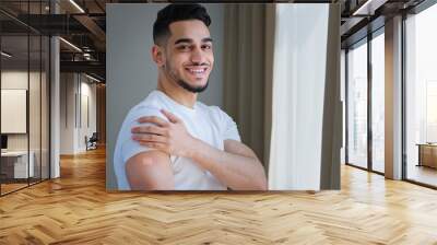 Happy satisfied indian guy arab man hispanic patient male posing indoors wears white t-shirt shows plaster on shoulder feels relief after vaccination injection with covid remedy medical immunization Wall mural