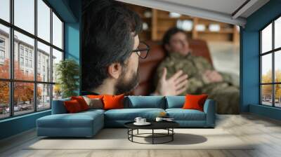 Middle aged Caucasian psychologist doctor man male speak talking to officer camouflage uniform military army soldier blurred patient guy lying on couch listening psychiatrist consultation session room Wall mural