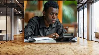 Portrait of an African young man sitting at home calculating his finance expenses with a calculator. He looks focused and determined as he prepares his budget and pays his bills. Wall mural