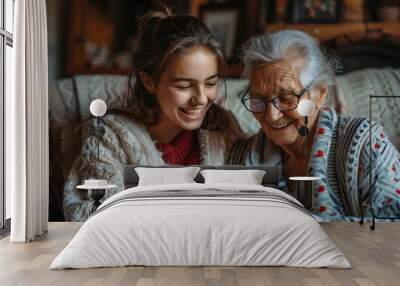 The beautiful young woman is smiling and laughing while teaching her beautiful grandmother, who is wearing eyeglasses, how to use a smartphone at home. Wall mural