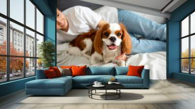 Young woman lying on the bed at home with Cavalier King Charles Spaniel dog and smiling. Wall mural