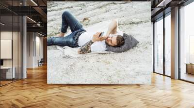 A young man on nature at the edge of the earth uses a smartphone. Lake, mountains. Weekend in nature with a gadget. Harmony with nature and technology. Wall mural