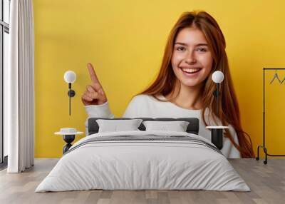 Smiling young woman in white sweater pointing finger up at copy space. Wall mural