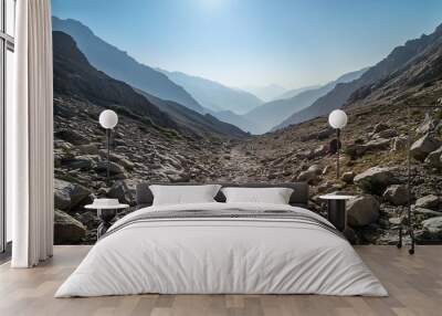 A rocky path leads up to the summit of a mountain range, with the sun shining brightly in the sky. Wall mural