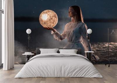 Beautiful young girl on a night beach with sand and stars holds the moon in her hands Wall mural