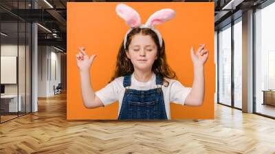 Close up portrait of carefree charming preschool girl kid cross fingers and closing eyes, wears pink bunny fluffy ears, isolated on orange background studio. Easter holiday and making wish concept Wall mural