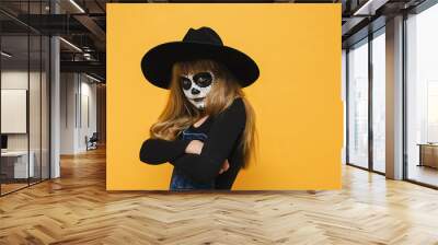 Portrait of angry sad little blonde girl child with Halloween makeup mask, wears black hat, crossed arms, unhappy looking to side, posing isolated over yellow studio background wall with copy space Wall mural