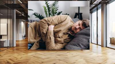 Sad young man lying on grey couch at home, suddenly feeling strong stomach ache, gastritis problem. Upset male squeezing belly with hands because of abdominal pain. Guy suffering from stomach ache Wall mural