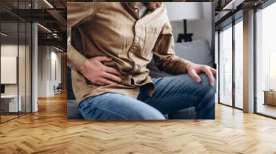 Sad young man sitting on couch at home, suddenly feeling strong stomach ache, gastritis problem. Upset male 30s squeezing belly with hand because of abdominal pain. Guy suffering from stomach ache Wall mural