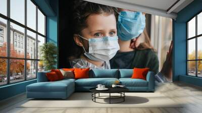 Unhappy young mother embracing upset little curly daughter with virus mask, sitting on windowsill at home, consoling sad preschool girl. Concept of coronavirus or COVID-19 pandemic disease symptoms Wall mural