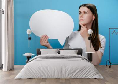 Worried pensive confused young female holding empty speech bubble, spreading arms put hand prop up on chin, dressed in white t-shirt, isolated on blue studio background wall. Emotion people concept Wall mural