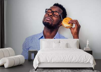 african american man enjoying the taste of hamburger.handsome and young afro man in a stylish shirt and glasses holding a burger on a white background.love of junk food diet Wall mural