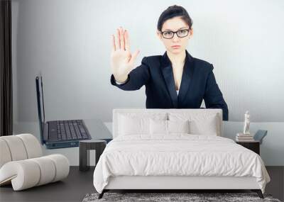 attractive strict brunette woman (business lady) in stylish business suit and glasses shows a hand gesture stop with laptop and a bunch of folders in the workplace office Wall mural