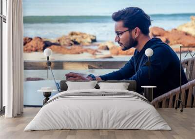 freelancing and remote work.handsome male student working laptop keyboard sitting in cafe on the beach with free internet.Young indian man using computer in summer vacation by the sea Wall mural