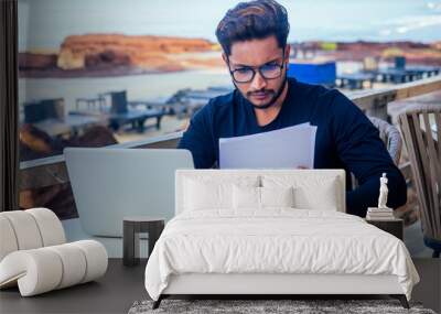 handsome and young successful indian man freelancing surfing remote work with a laptop on the beach by the ocean.india businessman freelance programming online copywriter paradise landscape dream job Wall mural