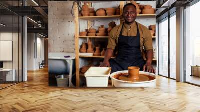 mixed race afro male potter with black apron and stylish dark shirt sitting at workshop table potter's wheel , applying glaze on handmade clay brown pot vase Wall mural