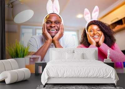 Young african american man and woman wearing cute easter bunny ears and painting eggs in the house Wall mural