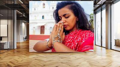 Young Indian woman in traditional sari red dress praying in a hindu temple goa india Hinduism.girl performing namaste gesture catholicism Delhi Street holi festival.om yoga meditation female model Wall mural