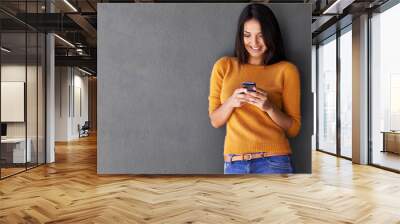 Bright and bubbly with some good news. An attractive young woman using a mobile phone while standing against a gray wall. Wall mural