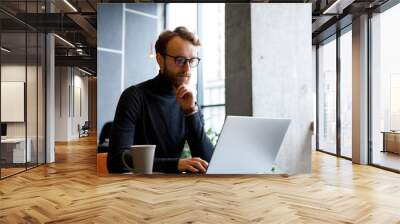 A young red-haired guy, a programmer or entrepreneur in glasses, works in a stylish cafe behind a laptop. Freelancer works remotely. Online communication. Small business. Wall mural