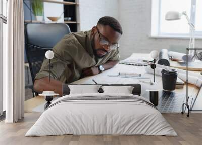 Afro-American architect working in office with blueprints.Engineer inspect architectural plan, sketching a construction project. Portrait of black handsome man sitting at workplace. Business concept. Wall mural