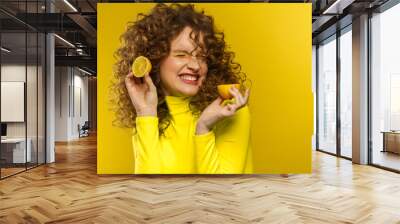 Young beautiful smiling women with curly hair tasting sour lemon halves emotionally. Funny girl with wavy hairstyle and bright make up isolated over yellow background. Copy space. Yellow lifestyle. Wall mural