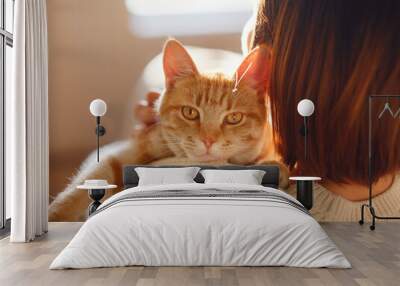 Young asian woman wears warm sweater resting with tabby cat on sofa at home one autumn day. Indoor shot of amazing lady holding ginger pet. Morning sleep time at home. Soft focus. Wall mural