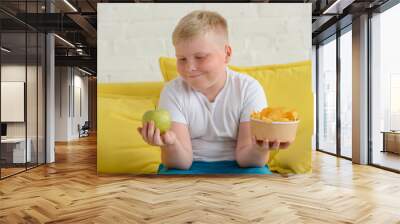 Happy fat boy looking at an apple and holding a cup of chips Wall mural