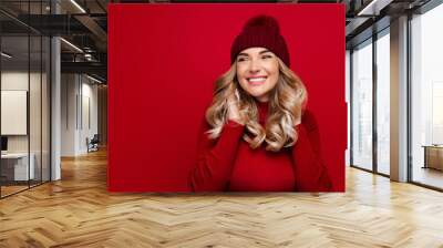 Charming smiling blonde woman touches curly hair with both hands, wears red wool turtleneck and hat, poses against red studio wall, enjoys the winter weather, looking at blank space for advertising Wall mural