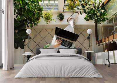 Freelancer woman working on laptop from home sitting at cozy balcony surrounded with green house plants. Businesswoman on remote occupation with computer at comfortable workplace near window. top view Wall mural