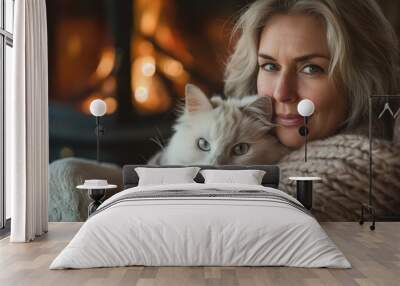 A woman cuddles with her fluffy white cat by the warm glow of a fireplace, wrapped in a cozy knitted blanket. Wall mural