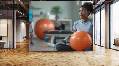 A woman holding an exercise ball in a fitness class, while others stand in the background, in a gym setting. Wall mural