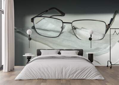 A medical mask lies on a white background, and dark-rimmed glasses are on the mask. Sunlight. Concept - protection against coronovirus, self-isolation mode Wall mural