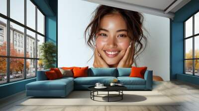 A close-up portrait of a young asian woman with long, dark hair, smiling brightly with her hands resting gently on her cheeks. Wall mural