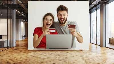 Happy smiling couple man and woman wearing grey and red t-shirt doing online shopping in front of laptop, holding credit cards on white wall background. Shopping of black friday and cyber monday. Wall mural