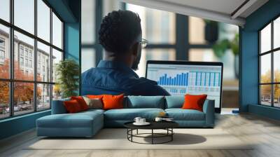 Businessman or financial accountant working with business spreadsheets on a laptop computer. African American man sitting at office desk and looking at screen of notebook PC, back view, over shoulder Wall mural