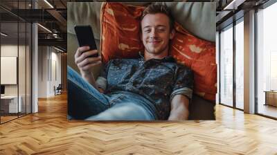 Relaxed young man in casual attire is happily lounging on a couch, using his smartphone and smiling while unwinding at home Wall mural