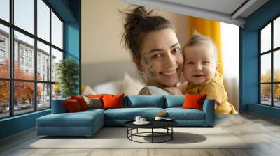 Smiling tattooed mother holding her happy baby indoors on a cozy morning, family love concept Wall mural