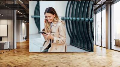 A stylish woman is sitting on a wall on city street and smiling at her phone. Wall mural