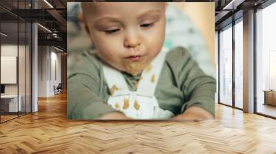 Baby eating with a face stained in food while in a child's dining chair Wall mural