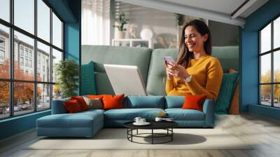 Portrait of a woman using laptop and a phone while sitting on a couch at home Wall mural