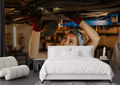 Professional female mechanic under lifted car in workshop examining car Wall mural