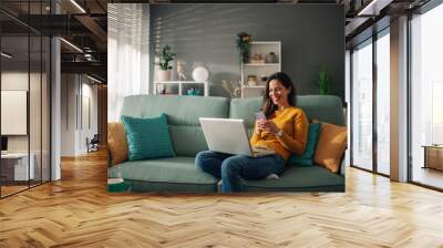 Woman using laptop and typing on a phone while sitting on couch at home Wall mural