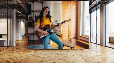 Young cheerful woman playing electrical guitar at home while dancing Wall mural