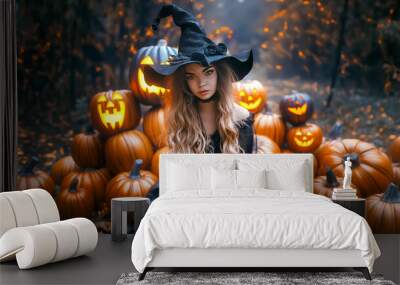 A young woman wearing a witch hat and dark dress sits amid carved pumpkins glowing in a forest filled with autumn leaves. The scene captures the essence of Halloween magic Wall mural