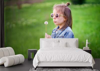 Beautiful little girl in denim clothes and rounded sunglasses blowing on a dandelion outdoor in the park Wall mural