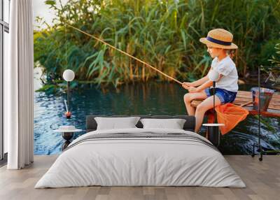 Little boy in straw hat sitting on the edge of a wooden dock and fishing in lake at sunset. Wall mural