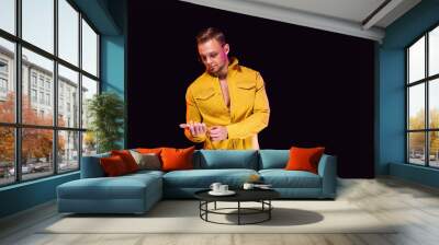 Positive young male looking away and adjusting trendy yellow shirt while standing under colorful light during party Wall mural